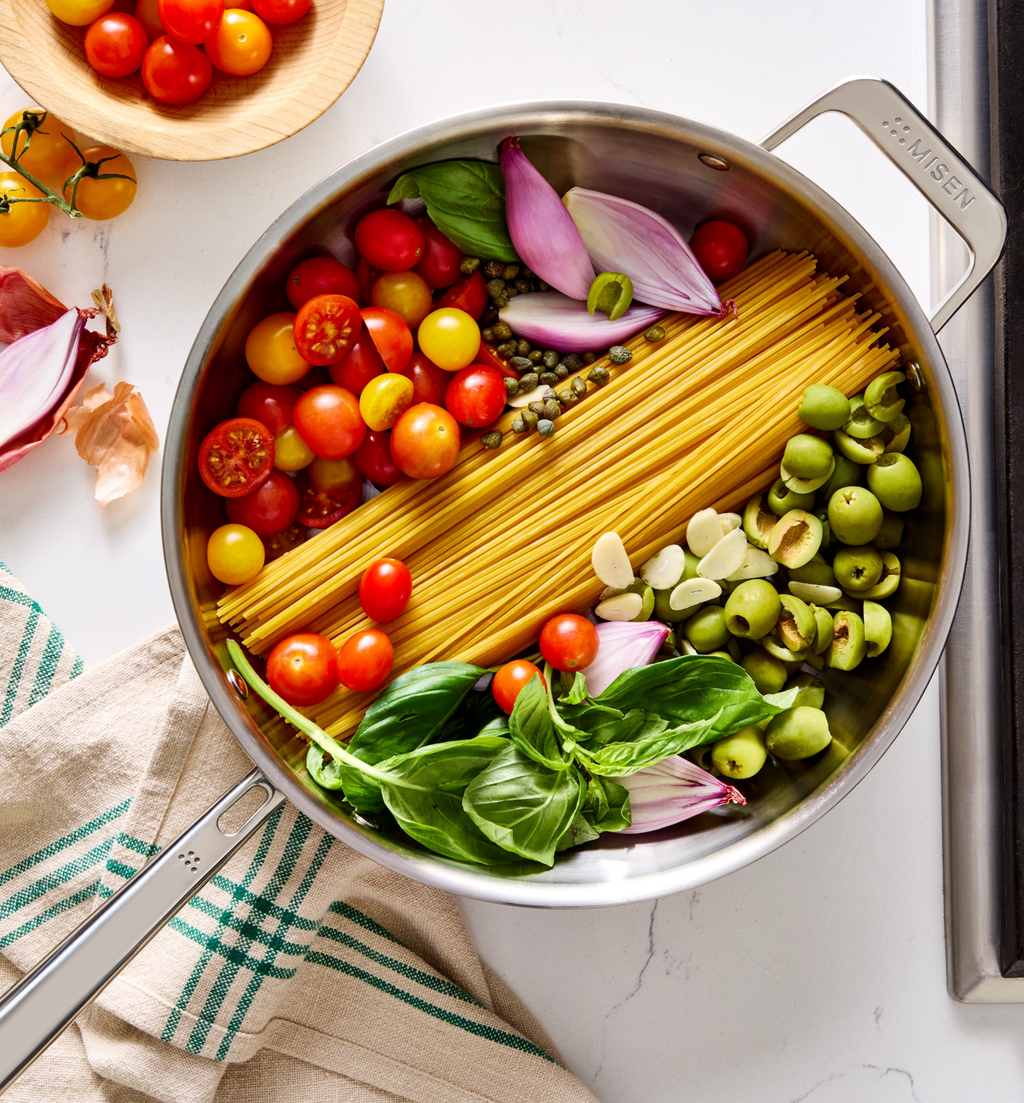 One-pot pasta ingredients in Misen stainless steel pan with spaghetti, cherry tomatoes, green olives, red onion, capers, garlic, and fresh basil